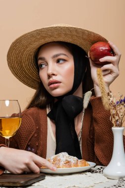 Stylish model holding apple near croissant and wine on table isolated on beige 