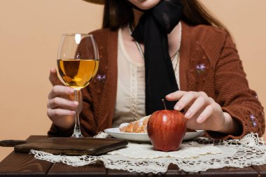 Cropped view of blurred woman touching glass of wine and apple on table isolated on beige 