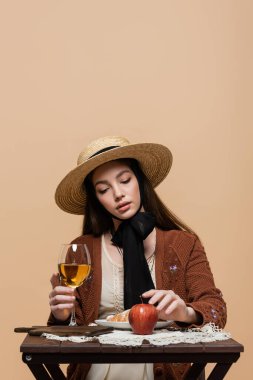 Fashionable woman in straw hat touching fresh apple and glass of wine isolated on beige 