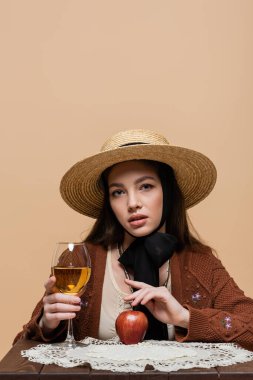 Stylish model in sun hat holding glass of wine and apple on table isolated on beige 