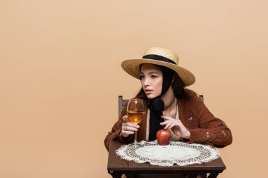 Young woman in sun hat touching wine glass and apple on table isolated on beige 