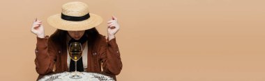 Trendy young woman in sun hat sitting near wine glass of table isolated on beige, banner 