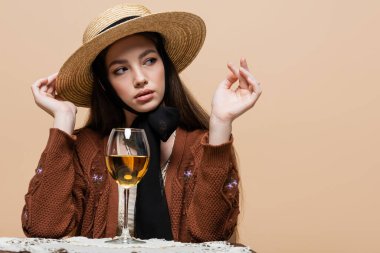Stylish woman in straw hat looking away near glass of wine on table isolated on beige 