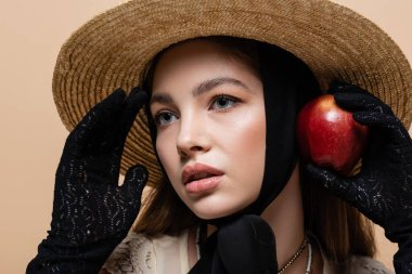 Portrait of pretty woman in gloves and straw hat holding apple isolated on beige 