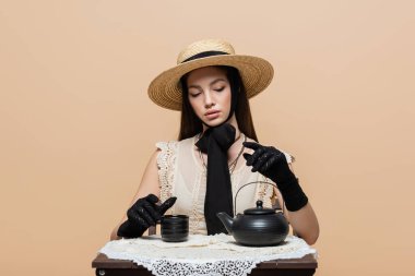 Stylish woman in straw hat and gloves sitting near teapot and cup isolated on beige 