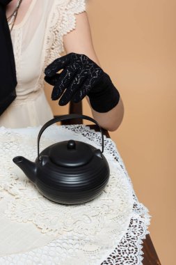 Cropped view of stylish woman in glove sitting near teapot on table isolated on beige  clipart