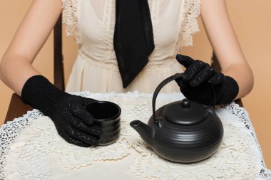 Cropped view of woman in gloves holding cup and teapot on table isolated on beige 