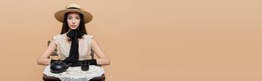 Fashionable woman in straw hat and gloves looking at camera near black teapot on table isolated on beige, banner 