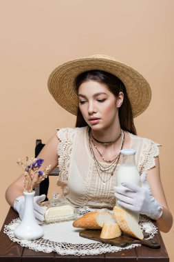 Trendy model in sun hat holding milk and butter near bread on table isolated on beige 