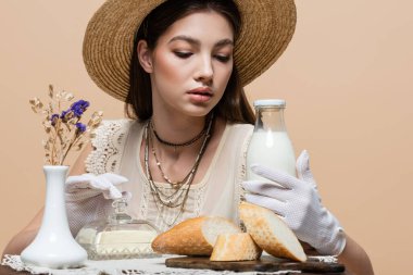 Trendy young woman in gloves touching bottle of milk near butter and bread on table isolated on beige 