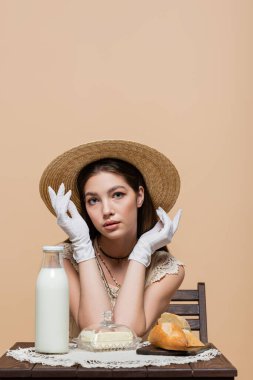 Pretty woman in sun hat and gloves looking at camera near bread and milk on table isolated on beige 