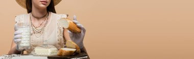 Cropped view of woman in gloves holding milk and bread near butter isolated on beige, banner 