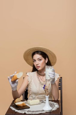 Trendy young woman holding bread and milk near butter and plants isolated on beige 