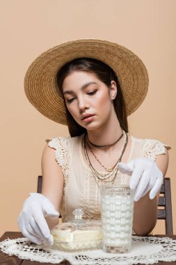 Trendy young woman in straw hat and gloves touching glass of milk and butter isolated on beige 