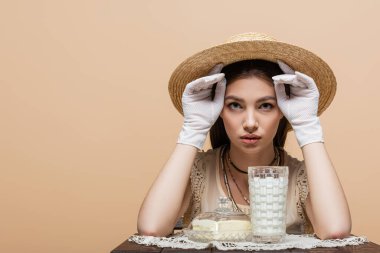 Brunette woman in sun hat and gloves looking at camera near butter and milk on table isolated on beige 