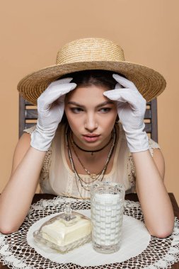 Pretty woman in sun hat looking away near milk and butter on table isolated on beige 