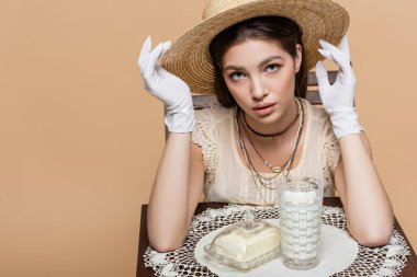 Stylish young woman in gloves looking at camera near milk and butter isolated on beige 