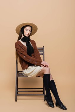 Young model in straw hat sitting on chair on beige background 
