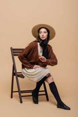 Pretty and stylish woman in sun hat pointing with finger while sitting on chair on beige background 