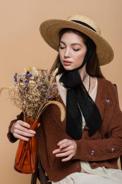 Trendy woman in sun hat looking at flowers in vase isolated on beige 