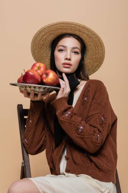 Portrait of stylish woman in straw hat holding apples on plate on beige background 