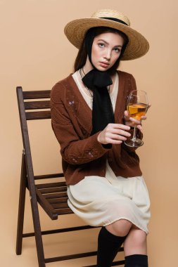Trendy model in sun hat holding glass of wine and sitting on chair on beige background 