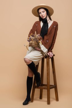 Fashionable woman in sun hat holding flowers while sitting on chair on beige background 