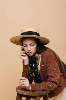 Trendy woman in straw hat and knitted cardigan looking at glass of wine on chair isolated on beige 
