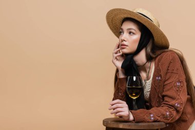 Fashionable model in straw hat looking away near glass of wine on chair isolated on beige 