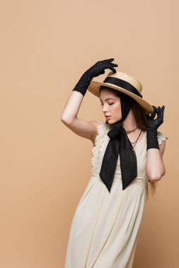 Stylish young woman in retro dress and gloves touching sun hat isolated on beige 