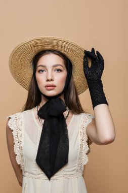 Brunette woman in straw hat and vintage dress looking at camera isolated on beige 
