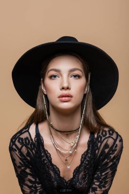 Portrait of trendy brunette woman in black hat looking at camera isolated on beige 
