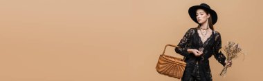 Young woman in black fedora hat holding wicker basket and dry flowers isolated on beige, banner 