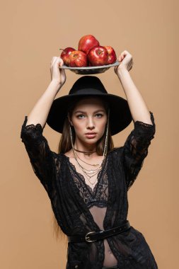 Model in fedora hat and guipure robe holding plate with apples above head isolated on beige 