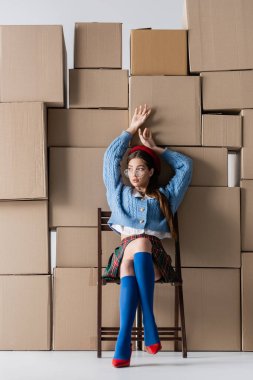 Trendy young woman n eyeglasses and beret sitting on chair near cardboard boxes on white background 