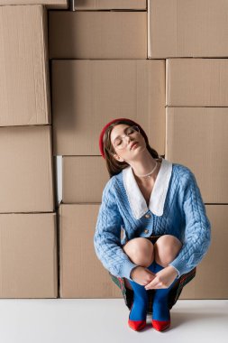 Stylish woman in eyeglasses and beret closing eyes near cardboard boxes on white background 