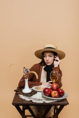 Stylish model in vintage clothes sitting near food and plants isolated on beige 