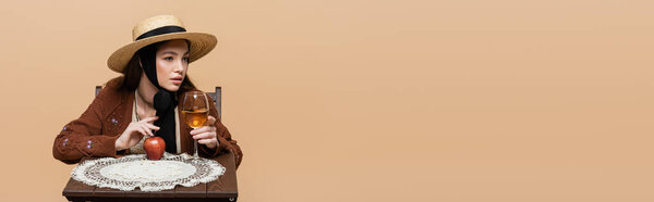 Стильная женщина в соломенной шляпе с вином возле яблока на деревянном столе, изолированном на бежевом, баннере 