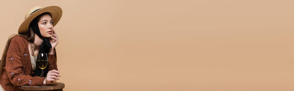 Стильная женщина в вязаном кардигане и соломенной шляпе, глядя в сторону бокала вина на стуле изолированы на бежевый, баннер 