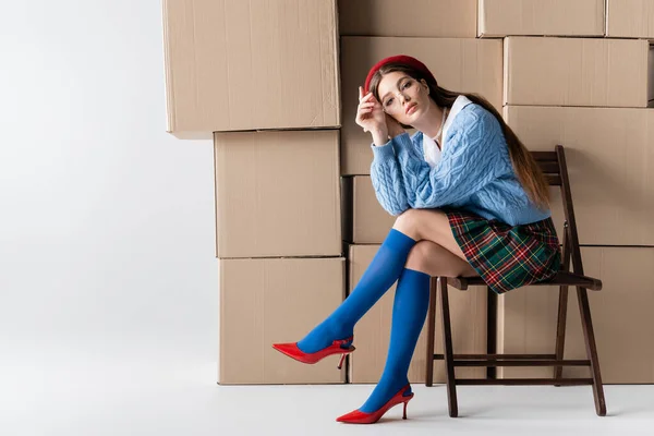 Trendy young woman in beret and skirt looking at camera while sitting on chair near cardboard boxes on white background