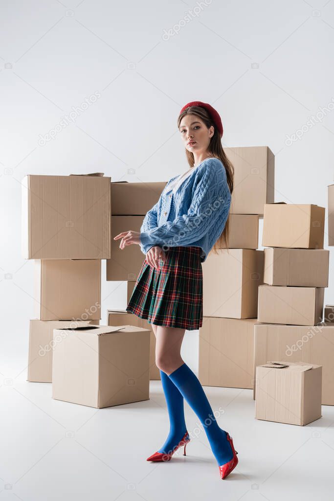 Full length of stylish model in heels and beret looking at camera near carton boxes on white background 