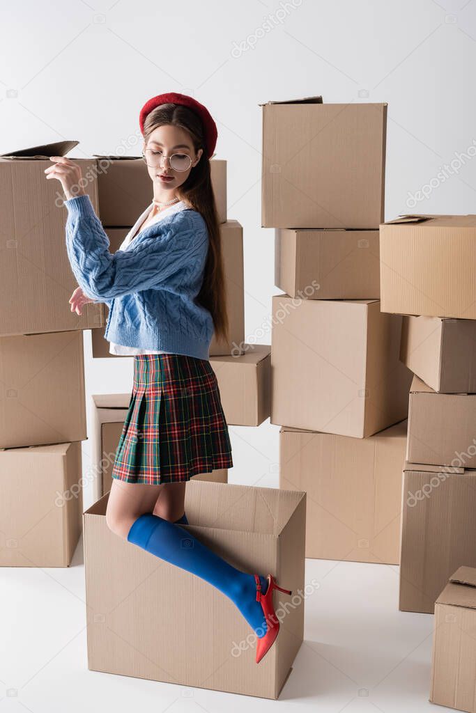 Trendy brunette woman in beret and skirt standing in box near cardboard packages on white background 