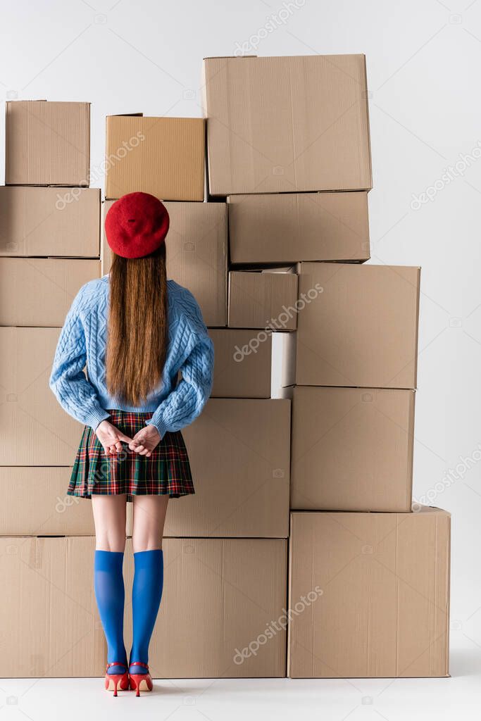 Back view of trendy young woman in beret and knee socks standing near cardboard boxes on white background 