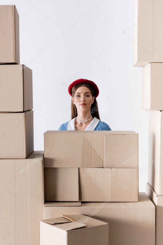 Trendy model in beret and eyeglasses looking at camera behind cardboard boxes isolated on white 