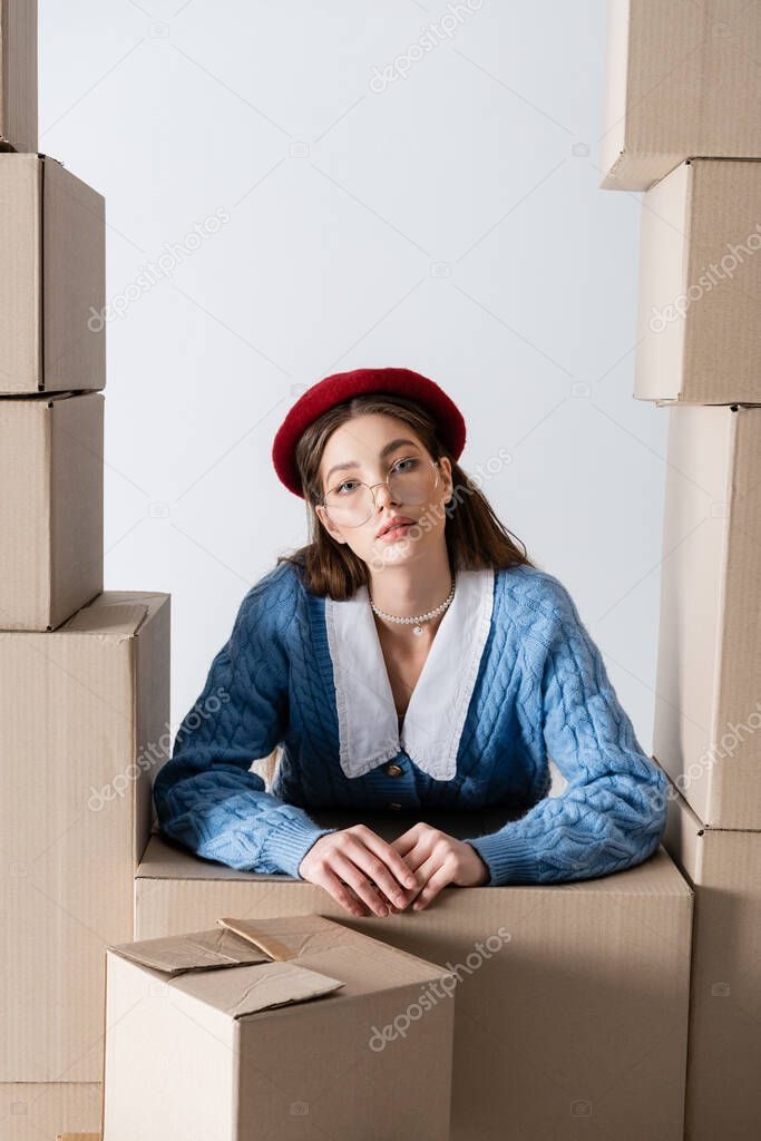 Stylish woman in beret looking at camera between cardboard boxes isolated on white 