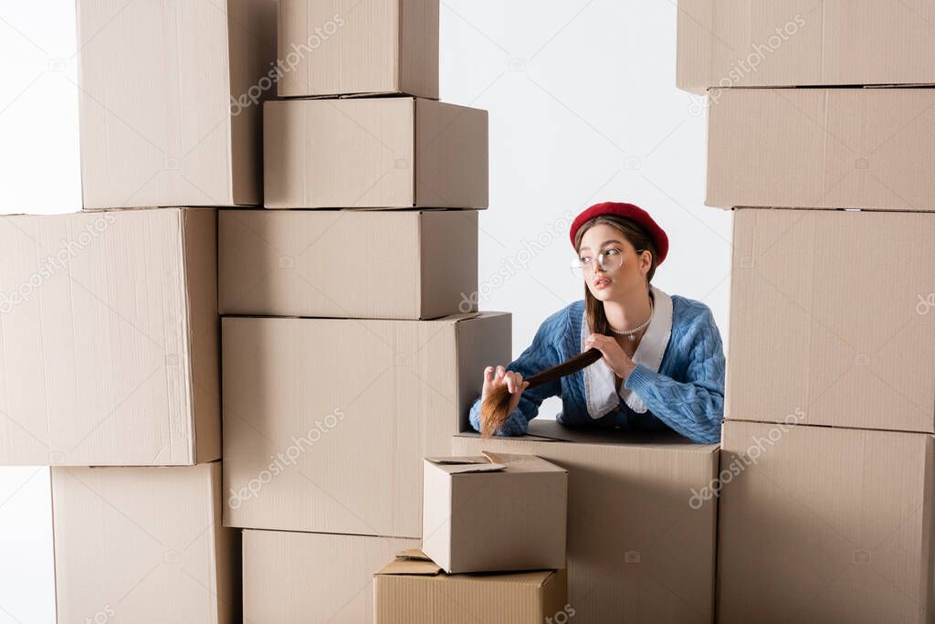 Trendy woman in eyeglasses and beret looking away between cardboard boxes isolated on white 