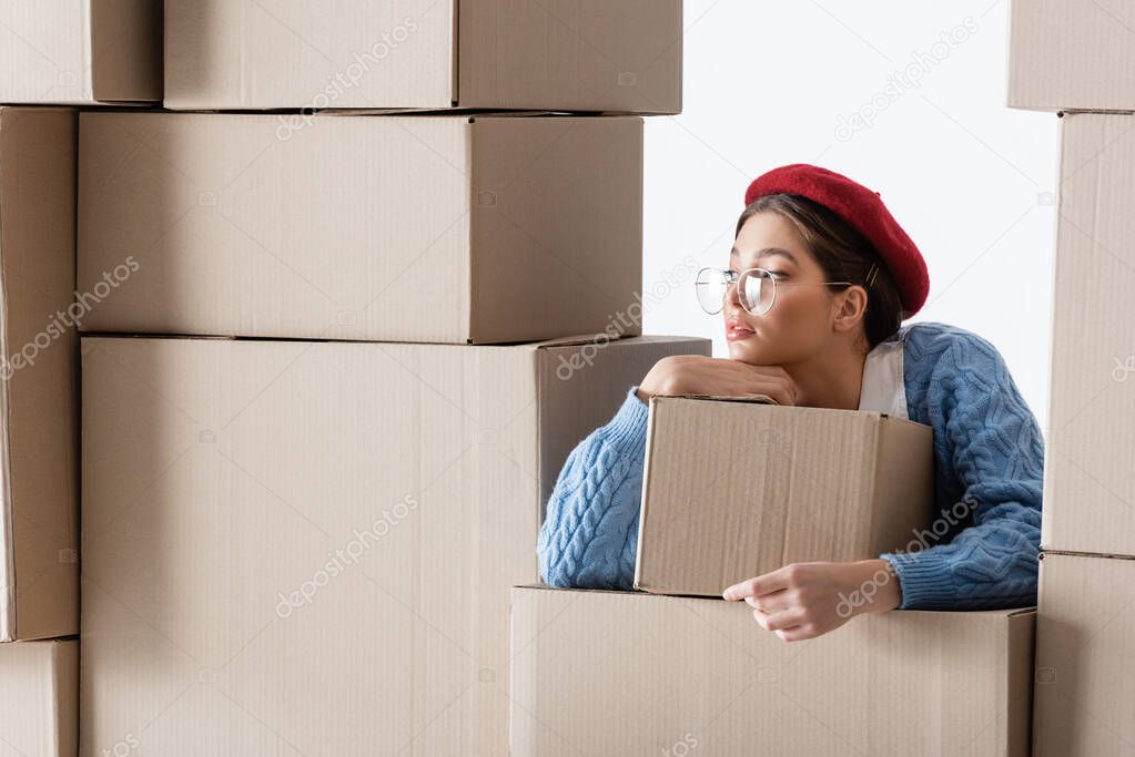 Trendy woman in beret and eyeglasses looking away near cardboard boxes isolated on white 