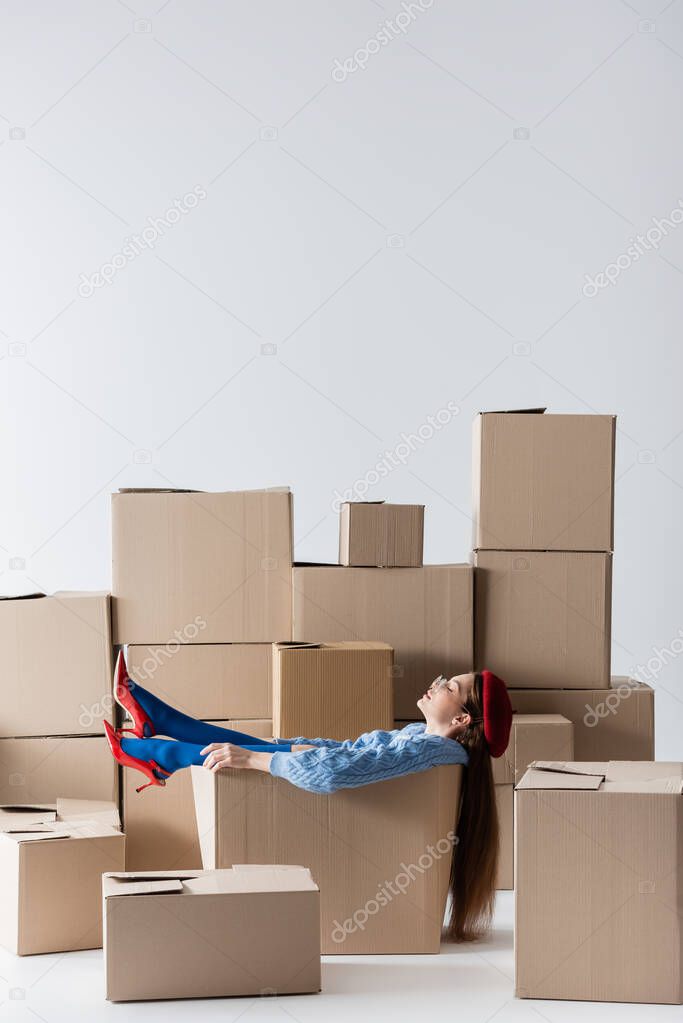 Side view of woman in heels and eyeglasses sitting in cardboard box on white background 