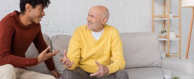Cheerful african american grandson talking to grandparent on couch at home, banner 