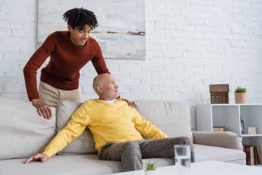 Smiling african american grandson standing near senior granddad on couch in living room  clipart